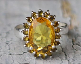 May include: A silver ring featuring a large, oval-cut yellow gemstone, surrounded by smaller, round yellow-brown stones. The ring's design is set against a textured, light-colored surface.