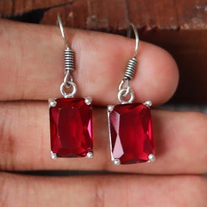 May include: A pair of drop earrings featuring rectangular, faceted deep red stones set in silver-toned metal. Each earring has a simple hook wire with a coiled silver detail at the top, suspending the vibrant red gemstone in a four-prong setting.
