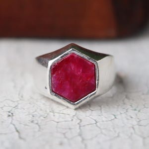 Könnte beinhalten: Ein Silberring mit einem großen, sechseckigen, rubinroten Stein. Der Ring hat ein dickes, poliertes Band, und der Edelstein ist ein leuchtendes Rot. Der Ring ist vor einem neutralen Hintergrund platziert.