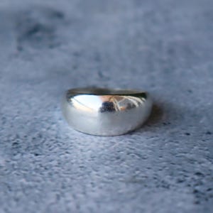 Puede incluir: Primer plano de un anillo plateado con un diseño redondeado y liso. El anillo refleja la luz, creando reflejos en su superficie. El anillo es una joya sencilla pero elegante.