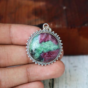 May include: Circular pendant with a silver-coloured beaded frame. The pendant features a polished stone with a mix of red and green colours. The stone is set in a silver-coloured metal setting.