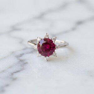 Peut inclure: Une bague argentée ornée d'une grande pierre ronde rouge foncé au centre. La pierre centrale est entourée d'un halo de huit petites pierres rondes transparentes disposées en étoile, sur un anneau en métal argenté lisse et poli.