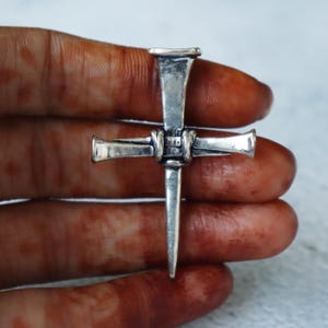 May include: A silver-colored cross pendant crafted from nails. The cross features a vertical nail with a pointed end and horizontal nails forming the arms. The nails are joined together with a wrapped design. The pendant is held in a hand.