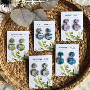 May include: A collection of polymer clay earrings in various colors, including green, blue, and purple, displayed on white cards within a woven basket. Each card reads "WRITTEN IN CLAY Polymer Clay Earrings". The earrings have a marbled effect.