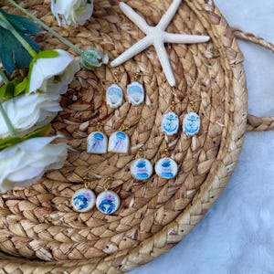 May include: Four pairs of dangle earrings featuring nautical and marine designs, including a blue crab, a jellyfish, a stack of stones, and an anchor. Each earring has a gold-tone metal hook and a white base with colorful illustrations.