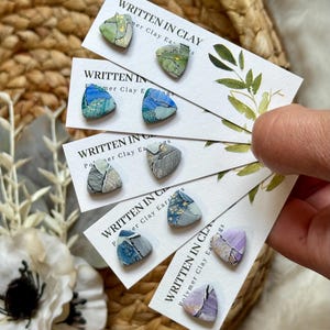 May include: A collection of polymer clay earrings in various colors, including green, blue, gray, and purple. Each earring is a unique triangular shape with abstract designs. The cards read "WRITTEN IN CLAY Polymer Clay Earrings."