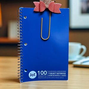 May include: A blue spiral-bound notebook with a gold paperclip and a pink bow-shaped embellishment. The notebook has the text "Jot 100 Sheets" and dimensions of 5.75 in x 8.75 in (14.6 cm x 22.2 cm).