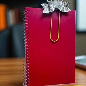 May include: A red spiral-bound notebook with a silver crown-shaped paperclip. The paperclip is attached to the top of the notebook and has a yellow paperclip. The notebook is on a wooden surface.