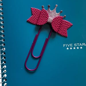 May include: A decorative purple paperclip with a pink bow and glittery silver crown sits on a teal notebook. The notebook has the text "Mead" and "FIVE STAR" printed on it.
