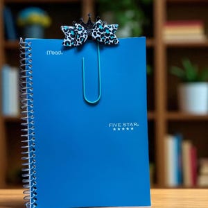 May include: A blue spiral notebook with the Mead and Five Star logos. A decorative paper clip with a bow and crown design is attached to the top of the notebook. The bow has a black and white leopard print with teal accents.