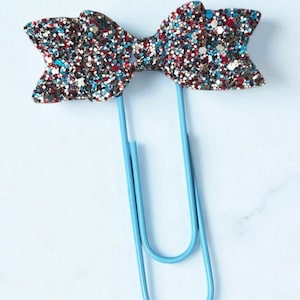 May include: A glittery bow-shaped paper clip with a mix of red, silver, and blue glitter. The bow is attached to a light blue metal paper clip. The background is a light, neutral color.