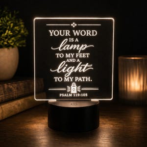 May include: A square, illuminated acrylic plaque featuring the engraved text: YOUR WORD IS A LAMP TO MY FEET AND A LIGHT TO MY PATH. PSALM 119:105. The design includes a small lantern icon and is mounted on a circular black base with a power button.