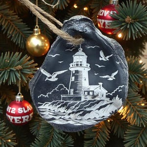 May include: A black oyster shell Christmas ornament featuring a white lighthouse scene with seagulls. The ornament hangs from a natural twine string and is displayed on a Christmas tree with other ornaments.