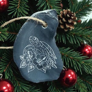 May include: A dark gray, irregularly shaped ornament with a white turtle and hibiscus flower design. The ornament hangs from a natural twine string and is displayed on a Christmas tree with red ornaments and a pine cone.