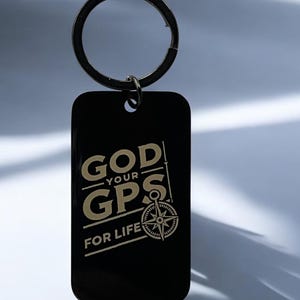 May include: Black rectangular keychain with a silver key ring. The keychain has the words "GOD - YOUR - GPS - FOR LIFE" in a gold color, along with a compass design. The keychain is a religious-themed accessory.