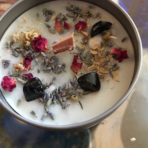 May include: A white candle with dried lavender, rose petals, and black stones embedded in the wax.