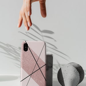 May include: A pink and black geometric phone case with glitter accents. The case is sitting on a gray concrete block and a gray concrete cylinder.