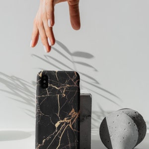 K&ouml;nnte beinhalten: Eine schwarze Handyh&uuml;lle mit einem goldenen Marmormuster. Die H&uuml;lle liegt auf einer wei&szlig;en Oberfl&auml;che mit einem grauen Betonblock und einem grauen Zylinder.