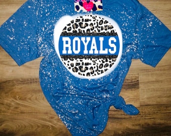 Puede incluir: Una camiseta azul con un diseño salpicado de lejía. La camiseta presenta un círculo con un fondo de estampado de leopardo y una franja azul con la palabra "ROYALS" en letras blancas.