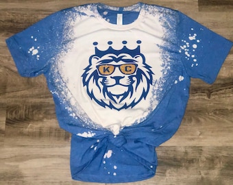 Puede incluir: Una camiseta azul con un diseño de lejía blanca de un león con gafas de sol y una corona. Las gafas de sol tienen las letras "KG" en purpurina dorada.