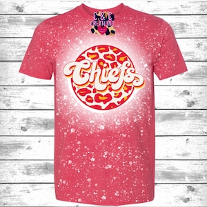 May include: Red t-shirt with a white bleach splattered design. The design features a circle with a leopard print pattern and the word "Chiefs" in a retro font.