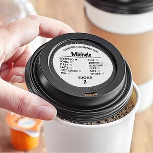 May include: A black coffee cup lid with the name "Michele" printed on it, along with a list of coffee options and sugar level. The lid is placed on a white paper coffee cup filled with coffee. The text "CAUTION CONTENTS HOT" is printed on the lid.