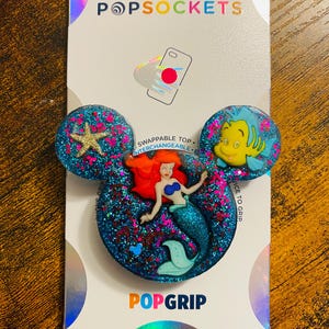 May include: A PopSockets PopGrip featuring a glittery blue design with a mermaid and a starfish. The PopGrip has a Mickey Mouse ear shape and includes the text "POPSOCKETS" and "POPGRIP".