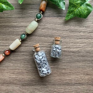 Könnte beinhalten: Zwei kleine Glasflaschen mit Korkstopfen, gefüllt mit silbernem Glitzer, auf einer Holzoberfläche. Eine Halskette mit grünen und orangefarbenen Steinen und Metallakzenten befindet sich im Hintergrund.