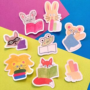 Puede incluir: Nueve pegatinas coloridas con animales leyendo libros. Las pegatinas están sobre un fondo azul y morado. Los animales incluyen un zorro, una rana, un mapache, un león, un conejo, un erizo y un perro.