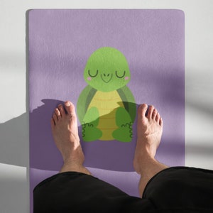 Op de afbeelding: Een paarse yogamat met een groene cartoon schildpad die erop mediteert. De blote voeten van een persoon bevinden zich aan weerszijden van de mat.