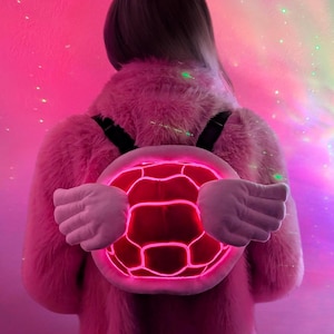 May include: A pink fuzzy jacket with a light-up turtle shell backpack. The shell has white wings and glows pink.