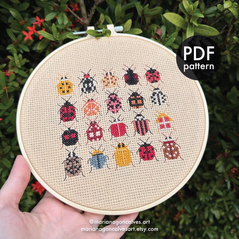 Peut inclure: Un cercle de broderie au point de croix beige avec un motif de coccinelles color&eacute;es. Les coccinelles pr&eacute;sentent diff&eacute;rents motifs et couleurs, notamment le rouge, le noir, le jaune et le rose. Le texte "PDF pattern" est dans un cercle noir. Le site web de l'artiste est &eacute;galement visible.