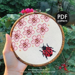 May include: A cross-stitch embroidery hoop with a floral and ladybug design. The pattern features pink flowers and a red and black ladybug on cream fabric. The round wooden hoop has a PDF pattern label. The design is for cross-stitch.