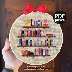 Pode incluir: Bordado em ponto cruz num bastidor, retratando uma estante com livros coloridos, gatos e um laço vermelho. O texto "PDF pattern" é visível. O design inclui o texto "@marianagoncalves.art" e "marianagoncalvesart.etsy.com".
