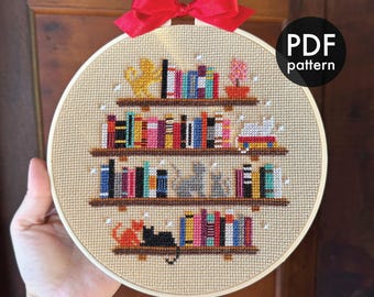 May include: A cross-stitch embroidery hoop featuring a bookshelf design with colorful books, cats, and a red bow. The text "PDF pattern" is visible. The design includes the text "@marianagoncalves.art" and "marianagoncalvesart.etsy.com".