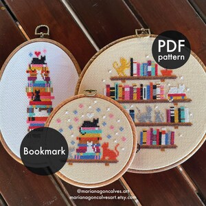 May include: Three cross-stitch embroidery hoops with cat and bookshelf designs. One hoop is a bookmark, another is a PDF pattern. The designs feature stacks of books, colorful spines, and black cats. The background is a light beige fabric.