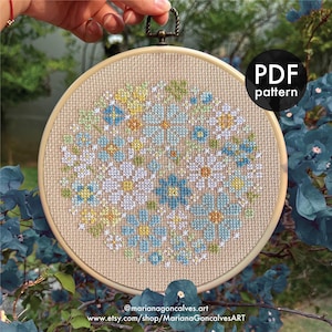 May include: A circular cross-stitch embroidery with a floral pattern in blue, yellow, green, and white. The text "PDF pattern" is visible. The design is set against a beige fabric background, held by a metal loop.