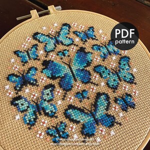 Peut inclure: Motif de point de croix avec des papillons bleus sur tissu beige, maintenu dans un cercle &agrave; broder en bois. Le motif pr&eacute;sente des papillons de diff&eacute;rentes tailles avec des d&eacute;tails d'ailes complexes. Le texte "PDF pattern" est dans un cercle noir. Les r&eacute;seaux sociaux de l'artiste sont &eacute;galement visibles.