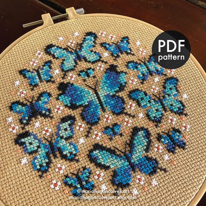 Peut inclure: Motif de point de croix avec des papillons bleus sur tissu beige, maintenu dans un cercle &agrave; broder en bois. Le motif pr&eacute;sente des papillons de diff&eacute;rentes tailles avec des d&eacute;tails d'ailes complexes. Le texte "PDF pattern" est dans un cercle noir. Les r&eacute;seaux sociaux de l'artiste sont &eacute;galement visibles.
