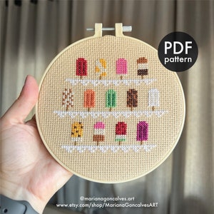 May include: A cross-stitch embroidery hoop featuring a design of colorful popsicle treats. The popsicles are in various colors, including red, orange, green, and brown. The text "PDF pattern" is in a black circle. The hoop is held by a hand.