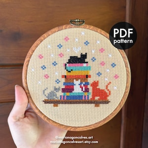 Puede incluir: Bordado de punto de cruz en un aro de madera que presenta una pila de libros coloridos con tres gatos. Un gato negro está sentado encima de los libros, con un gato gris y naranja a cada lado. El texto "PDF pattern" está en un círculo negro.