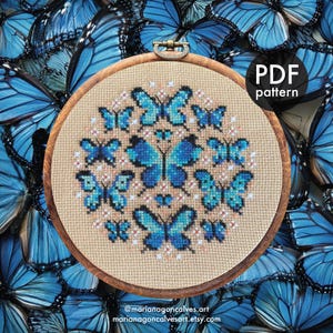 Peut inclure: Un cercle &agrave; broder en bois avec un motif de broderie au point de croix de papillons bleus. Le texte "PDF pattern" est visible. L'arri&egrave;re-plan est un collage d'ailes de papillons bleus. Informations de la boutique Etsy de l'artiste.