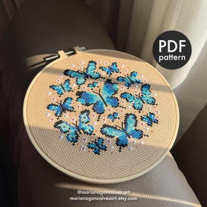 Peut inclure: Un cercle de broderie au point de croix avec un motif de papillons bleus et de petites fleurs blanches sur un tissu beige. Le texte "PDF pattern" est dans un cercle noir. Le nom et l'URL de la boutique Etsy sont en bas.