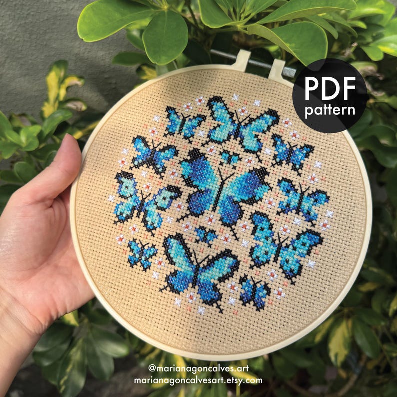 Peut inclure: Un cercle de broderie au point de croix avec un motif de papillons bleus et de petites fleurs blanches sur un tissu beige. Le texte "PDF pattern" est dans un cercle noir. Le texte "@marianagoncalves.art marianagoncalvesart.etsy.com" est en bas.