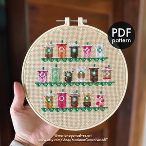 May include: A cross-stitch embroidery hoop with a design of coffee cups in various colours. The pattern includes green accents and the text "PDF pattern". The Etsy shop URL is also visible, with the shop name and website address.