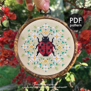May include: A cross-stitch embroidery of a red ladybird with black spots and a black head, encircled by white, yellow, and green floral details. The design is set in a round wooden hoop. The text "PDF pattern" is visible. The artist's name and shop URL are also displayed.