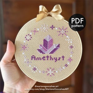 Könnte beinhalten: Ein Kreuzstichmuster mit einem violetten Amethystkristall mit weißem Umriss. Der Kristall ist von einem Kranz aus weißen Schneeflocken umgeben und das Wort "Amethyst" ist unter dem Kristall gestickt.