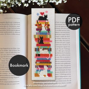 Pode incluir: Um marcador de livros feito à mão com um design de ponto cruz. O marcador de livros apresenta uma pilha de figuras coloridas, incluindo um gato, um cão e uma pessoa, com um coração no topo. A palavra "Bookmark" é visível.