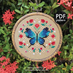 Op de afbeelding: Een ronde kruissteek borduurring met een blauwe vlinder met zwarte accenten, omringd door rode en witte bloemen. Het ontwerp is geplaatst op een beige stoffen achtergrond, omlijst door een houten ring. De tekst "PDF pattern" is zichtbaar.