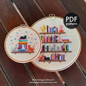 Può includere: Due schemi a punto croce in cerchi di legno. Uno raffigura una pila di libri con gatti, l'altro scaffali con libri e gatti. Il testo "PDF pattern" è visibile. Lo sfondo è in tessuto beige chiaro.